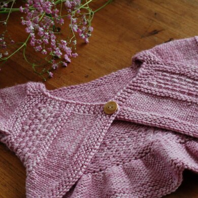 A pink cardigan with a single wooden button lays on an oak table, with a sprig of pink baby's breath above the neck opening. The cardi has bands of texture running diagonally from neck to armpit and a curved bottom edge